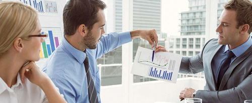 A young businessman, with a woman sitting next to him, shows another man a printed chart