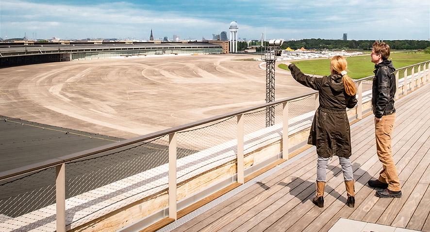 A young woman stands with a young man on the visitor terrace of Tempelhof Airport and points with her hand to the former airport grounds 